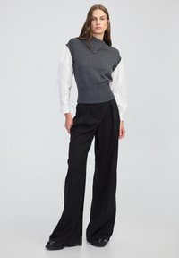 Grijze gebreide mouwloze top met een hoge kraag, gecombineerd met zwarte wijde broek en zwarte schoenen, gepresenteerd tegen een effen achtergrond.