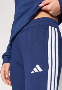 Μπλε σκούρο παντελόνι φόρμας με ελαστική μέση, που διαθέτει τρεις λευκές ρίγες κατά μήκος των πλαϊνών και το λογότυπο της Adidas στο αριστερό μηρό.