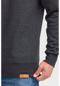 Bluza gri cu textură reiată, cu o etichetă din piele la tiv. Mânecile și banda de la talie sunt, de asemenea, reiate pentru o potrivire strânsă.