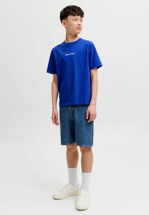 Chlapec stojící, oblečený v modrém tričku, džínových šortkách, bílých ponožkách a bílých teniskách, hledící doprava.