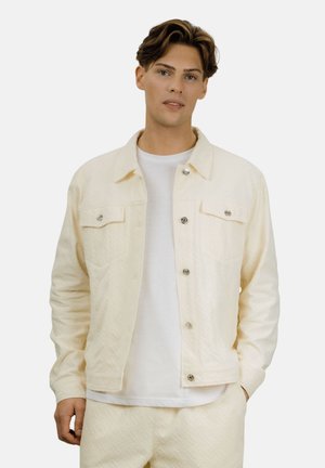 Ung mann iført en kremfarget, teksturert jakke over en hvit T-skjorte og matchende kremfargede bukser, stående foran en ensfarget bakgrunn.