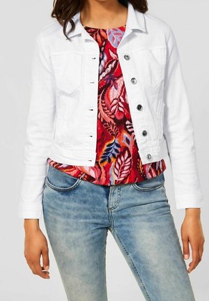 Kvinde iført lyseblå jeans, en farverig rød blomster-top og en åben hvid denimjakke med sølvknapper.