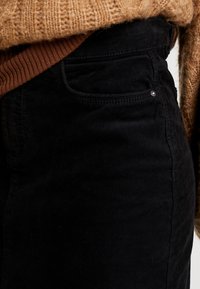 Zwarte corduroy broek met een gestructureerd oppervlak, voorzien van een zijzak en een lichte glinstering van een metalen knoop op de tailleband.