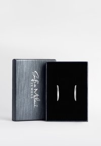 Silverfärgade örhängen i form av hoopar som visas i en svart sammetssydd låda med en strukturerad grå utsida. Varumärket präglat i vitt på locket.