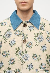 Άνδρας που φοράει μπεζ πουκάμισο με μπλε λουλουδένιο σχέδιο και μεγάλο μπλε γιακά, κουμπωμένο στην κορυφή με δύο σκούρα κουμπιά ορατά.