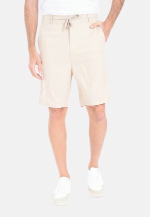 Uomo che indossa pantaloncini beige con coulisse, scarpe da ginnastica bianche e una camicia bianca, in piedi con una mano in tasca su sfondo bianco.