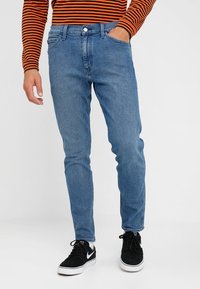 Blå denimjeans med slim fit och fem fickor. Har en enda knappstängning och lätt blekning. Bärs med svarta sneakers.