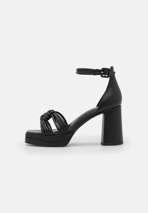 Sort højhælet sandal med ankelrem, knyttede frontstropper og chunky blokhæl på hvid baggrund.