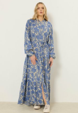 Donna in piedi contro il muro che indossa un maxi abito a fiori beige e blu con bottoni, maniche lunghe e sneakers bianche.