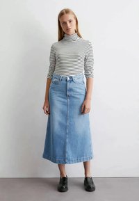 Kvinna i svart-vit randig polotröja, ljusblå denim midi kjol och svarta loafers som står mot en vit vägg.
