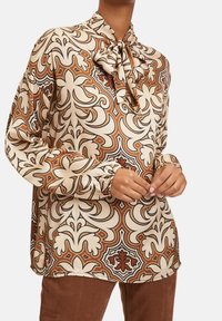 Långärmad blus i beige satin med bruna, orange och svarta blommönster. Har en knytkrage och detaljer med rynkade manschetter.