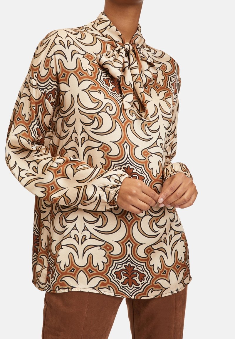Långärmad blus i beige satin med bruna, orange och svarta blommönster. Har en knytkrage och detaljer med rynkade manschetter.