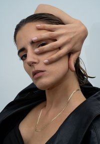 Nahaufnahme einer Person mit einer schwarzen Lederjacke, die eine goldene Kettenhalskette trägt, teilweise verdecktem Gesicht und glatter Haut mit neutralem Nagellack.