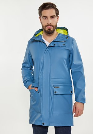 Bearded man wearing blue waterproof hooded jacket with yellow lining, hands in front pockets, standing against plain background.