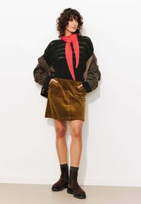 Bottines en suede marron, jupe en velours côtelé olive avec poches, pull à rayures noires superposé, et un foulard rouge au cou. Fond minimaliste.