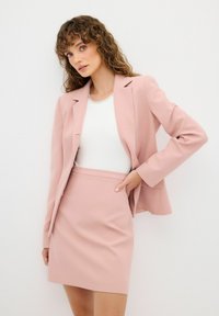Blazer de corte entallado en rosa claro y falda mini a juego, con un diseño estructurado y tejido suave, llevados sobre una blusa blanca con escote joya.