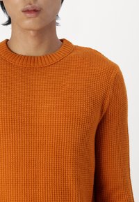 Pomarańczowy sweter wykonany na drutach, z prążkowanym, okrągłym dekoltem, fakturowanym splotem i dopasowanymi rękawami. Bez widocznych wzorów ani ozdób.