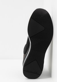 Semelle de basket noire avec motifs géométriques et marquage "Michael Kors" sur une surface blanche.