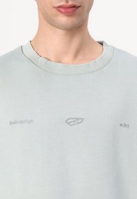 Light gray crewneck sweatshirt with ribbed neckline. Features embossed logo and text details, offering a smooth, soft texture.