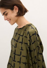 Top verde oliva realizzato in tessuto liscio, caratterizzato da un audace motivo a griglia nera. Il design presenta un collo ampio e una vestibilità morbida.