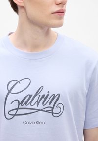 Jasnoniebieska bawełniana koszulka z krótkim rękawem, z wyraźnym ciemnoszarym nadrukiem "Calvin Klein" na klatce piersiowej.