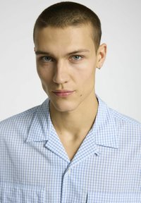 Šviesiai mėlyna trumpų rankovių marškinėlių su tinklelio raštu, turinti stovyklos apykaklę ir priekinį kišenę. Medžiaga atrodo lygi ir lengva.