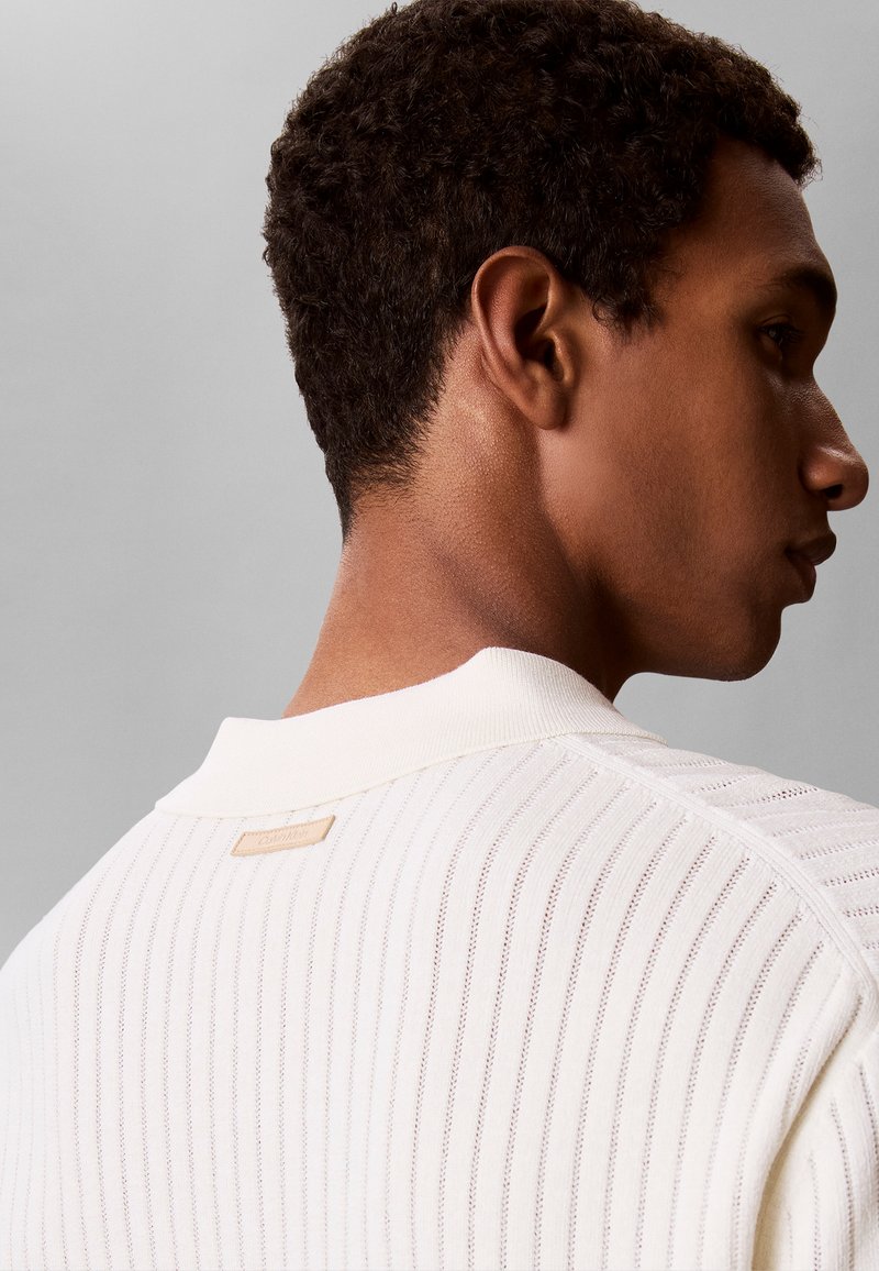 Gros plan sur le cou et l’épaule d’un homme portant un pull en maille blanche texturée avec une petite étiquette beige au niveau du col arrière.