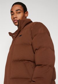 Giacca puffer marrone con colletto alto, caratterizzata da un logo Nike nero sul petto, texture a coste e silhouette oversize.