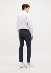 Camisetta bianca a maniche lunghe con leggero lucido, abbinata a pantaloni slim-fit blu scuro. Il modello indossa scarpe da ginnastica bianche e calzini chiari.