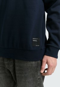 Marinblå sweatshirt i ett mjukt tyg med ribbad midja, som har en fyrkantig svart etikett på vänster sida nära fållen och långa ärmar.