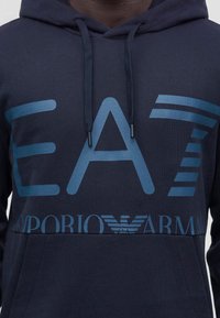 Sweat à capuche bleu foncé avec un grand logo bleu clair "EA7 Emporio Armani" sur la poitrine et des cordons noirs sur la capuche.