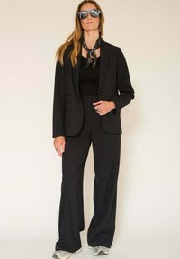 Costume noir à rayures avec un blazer ajusté et un pantalon large. Assorti avec un haut noir et une écharpe à motifs, complété par des lunettes de soleil réfléchissantes.