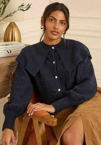 Námornícky pletený kardigán s golierom, s vzorom copánkov a striebornými gombíkmi. Textúrované rukávy a rebrované manžety, nosený cez hnedú sukňu.