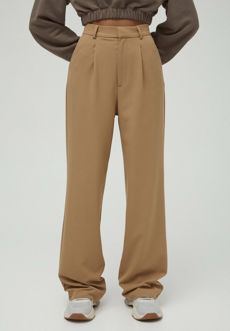 Pantalon beige taille haute, jambes larges, associé à des baskets blanches, porté par une personne les bras posés sur les hanches sur un fond clair uni.