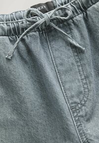 Ljusblå denimbyxor med elastisk midja och knytband, som visar framfickan och inre varumärkesetikett.