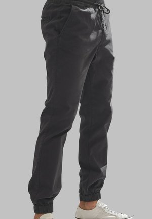 Pantalon de jogging noir avec taille et poignets élastiques, porté avec des baskets blanches à lacets, montré de la taille aux chevilles sur fond gris.