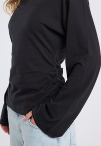 Black long-sleeve top made of soft fabric with a ruched side detail and flared sleeves, paired with light blue denim jeans.