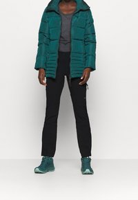 Petrolinsininen toppatakki vaakasuorilla ompeleilla, alla harmaa paita, mustat housut ja petrolinsiniset vaelluskengät. Vetoketju ja kiristimet näkyvissä.