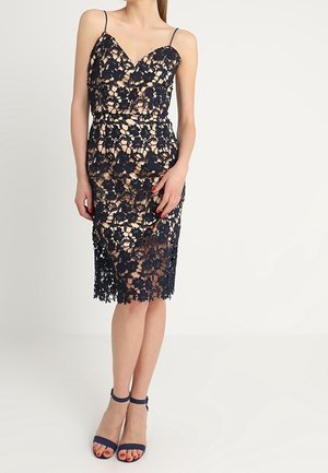 Kobieta w beżowej sukience bez rękawów, do kolan, z czarną koronką w kwiatowy wzór oraz czarnymi szpilkami z paskiem wokół kostki, stojąca na tle jasnej, jednolitej ściany.