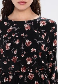 Černá květinová blůza s kulatým výstřihem, dlouhými rukávy a jemným vzorem bílých a burgundských květů na měkké tkanině.