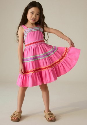 Fiatal lány rózsaszín ujjatlan ruhában, színes hullámos csíkokkal, kibontva a szoknyát, neutrális háttér előtt áll.