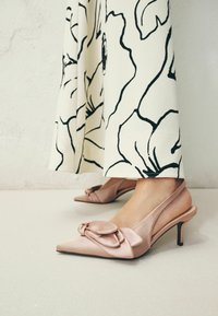 Femme portant des escarpins slingback rose clair à bout pointu avec des nœuds, assortis à un pantalon blanc à jambes larges à imprimé floral noir audacieux.