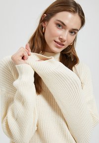 Roomkleurige gebreide trui met een hoge hals, lange mouwen en een textuurpatroon. Opvallende manchetdetails met oversized mouwen.