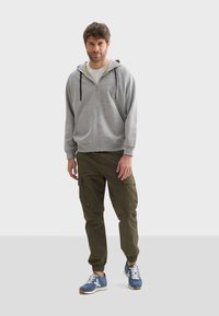 Un sweat à capuche gris zippé avec des cordons noirs, un t-shirt blanc en dessous, un pantalon cargo olive avec des poches, et des baskets bleues avec des lacets blancs.
