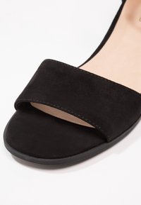 Sandale en daim noir avec une large sangle sur le dessus, semelle plate et détails de couture subtils le long des bords. Semelle intérieure beige lisse visible.