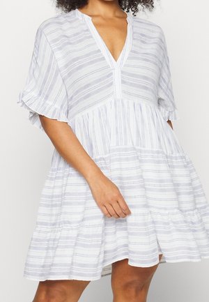 Woman in a white and light gray striped short-sleeve V-neck dress with ruffled tiers and hem, hand resting near waist.