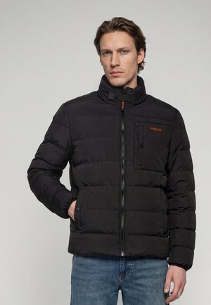 Homme portant une veste matelassée noire avec une fermeture éclair et des poches à boutons, debout devant un fond gris uni.
