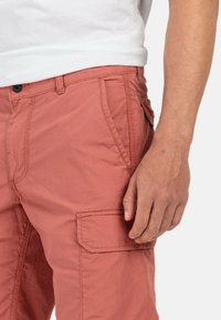 Mann in korallenfarbenen Cargo-Shorts und einem weißen Hemd, mit der Hand entspannt an der Seite vor einem schlichten Hintergrund.