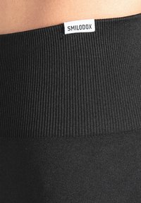 Schwarzer elastischer Bund mit gerippter Textur, der ein weißes Etikett mit der Aufschrift "SMILODOX" trägt. Das Material wirkt glatt und dehnbar.