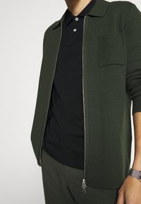 Mörkgrön cardigan med hela blixtlås, krage, strukturerad tyg och bröstficka, lager på en svart polo.